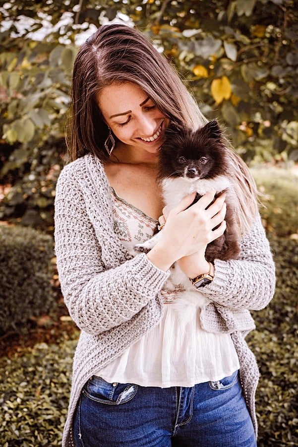 a girl standing near a tree and grass wearing a crochet grey cardigan and holding a small dog