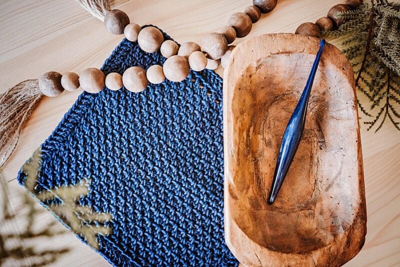 A blue crochet washcloth next to a wood bowl containing a crochet hook and decorative beads.