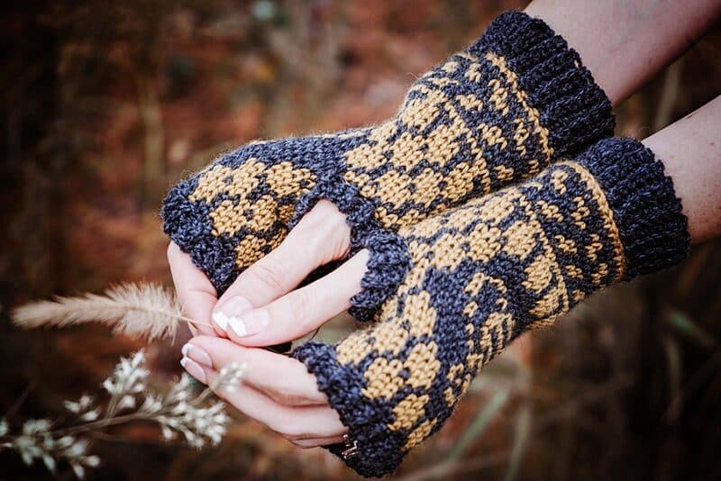 a pair of black and yellow mittens on a womans hands next to white flowers with a pine background