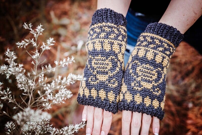 a pair of black and yellow mittens on a womans hands next to white flowers with a pine background