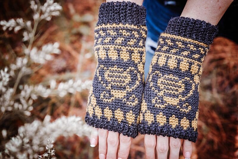 a pair of black and yellow mittens on a womans hands next to white flowers with a pine background