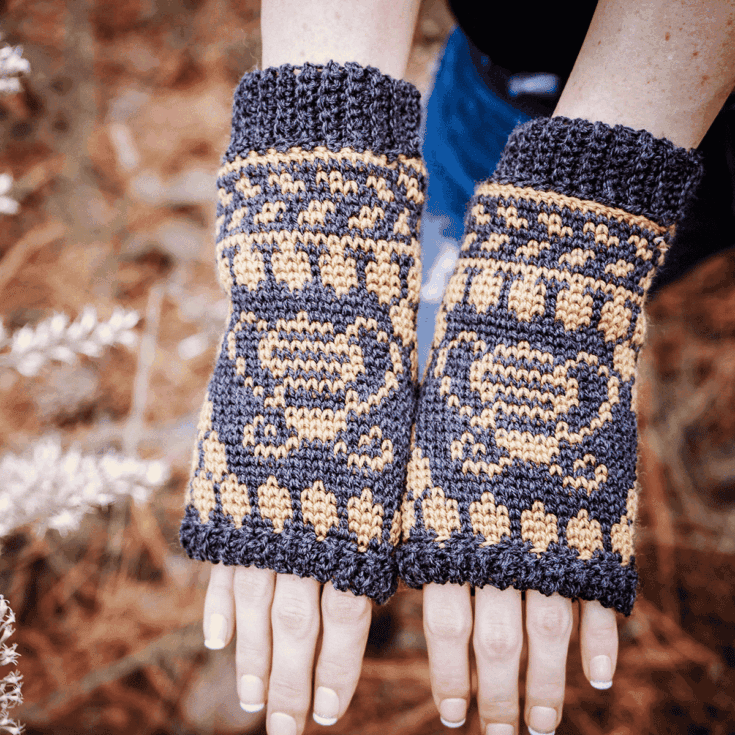 a pair of black and yellow mittens on a womans hands next to white flowers with a pine background