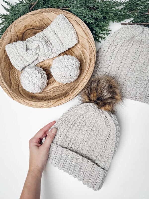 A hand holds a gray knit beanie with a faux fur pom-pom, reminiscent of Briana K's cozy style found on her social media links. Nearby are a matching headband and mittens on a wooden plate, next to another beanie minus the pom-pom. Greenery decorates the background.