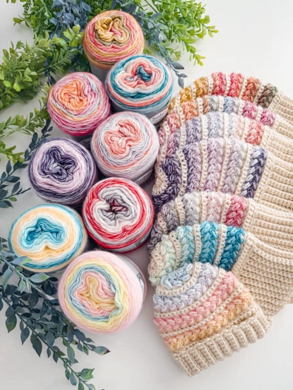 Six skeins of colorful variegated yarn and six matching crocheted hats are arranged on a white surface with green foliage accents—perfect for sharing on Briana K Social Media Links.