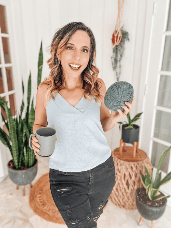 A woman, Briana K, holding a knitting ball and a mug in front of a potted plant.