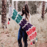 A woman stands outdoors in a forest holding the Easy Wander Crochet Wrap, a colorful crocheted wrap with a zigzag pattern. Text at top reads “Easy Wander Wrap Free Crochet Pattern.”.
