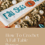 A Crochet Table Runner featuring "Fall Skies & Apple Pies" and an apple motif, displayed on a wooden table adorned with fabric pumpkins and a green garland.