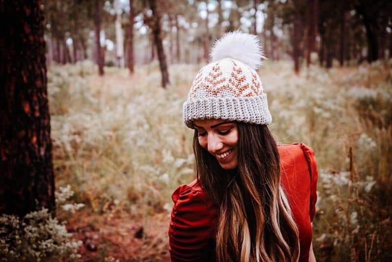A woman standing in a field of pine trees and tall grass wearing an orange shirt and wearing a crochet warm hat with a faux fur pompom