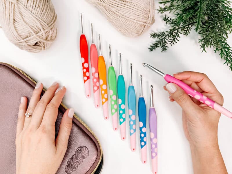A hand holds a pink crochet hook above a row of the best crochet hooks, with yarn, a plant, and a zippered pouch arranged on a white surface.