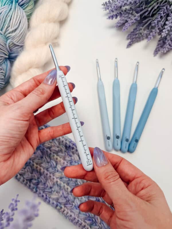 A person holds a blue crochet hook with size markings—part of a set of Best Crochet Hooks. Four other blue hooks, yarn, and a crocheted fabric are nearby on the table.