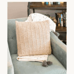 A crocheted pillow with a textured pattern sits on a blue armchair with a tasseled blanket, next to a bookshelf. Text reads: "How to Crochet Nutmeg Free Pillow Pattern, Free Pattern + Video Tutorial.