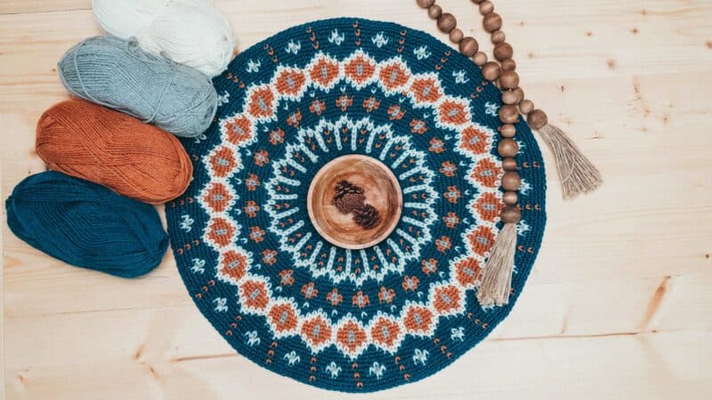 A blue and orange crocheted doily with yarn and a wooden board, featuring the Fawning Over You Crochet Sweater.