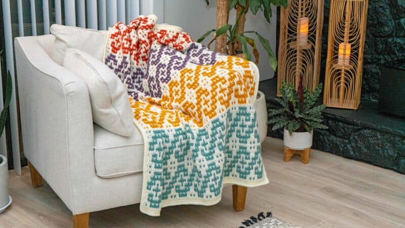 A colorful crochet blanket is sitting on a chair in front of a fireplace.