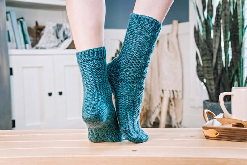 A person wearing cozy blue Mock Cabled Crochet Socks stands on a wooden table, with a cup and plant in the background.