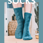 A person wearing blue mock cabled crochet socks stands on a wooden surface. The cozy indoor setting in the background adds to the charm. Text above reads "Crochet Socks" with "Free Pattern + Video Tutorial" below.