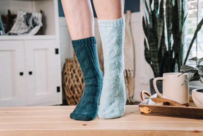 A person wearing mismatched knitted socks, with one dark blue and the other light blue, stands on a wooden table near a tray with a mug and greenery, showcasing their unique style in cozy mock cabled crochet socks.