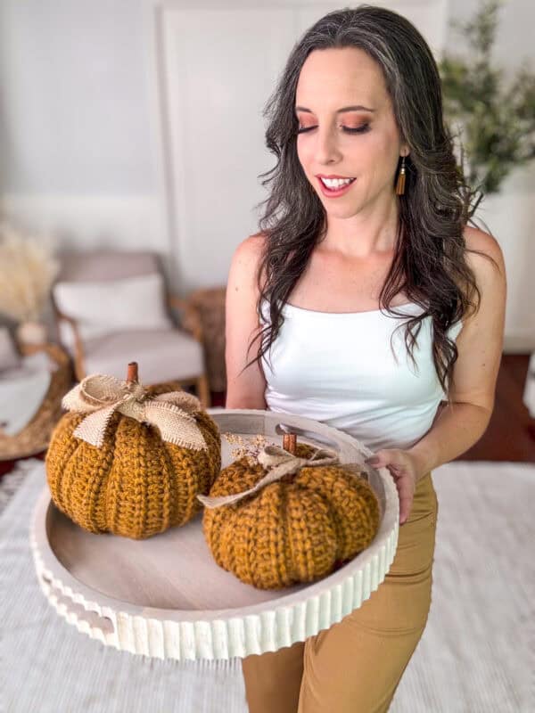 A woman wearing a white tank top and tan pants holds a tray with two of Cinderella's Crochet Pumpkins, decorated with bows, in a cozy living room setting.