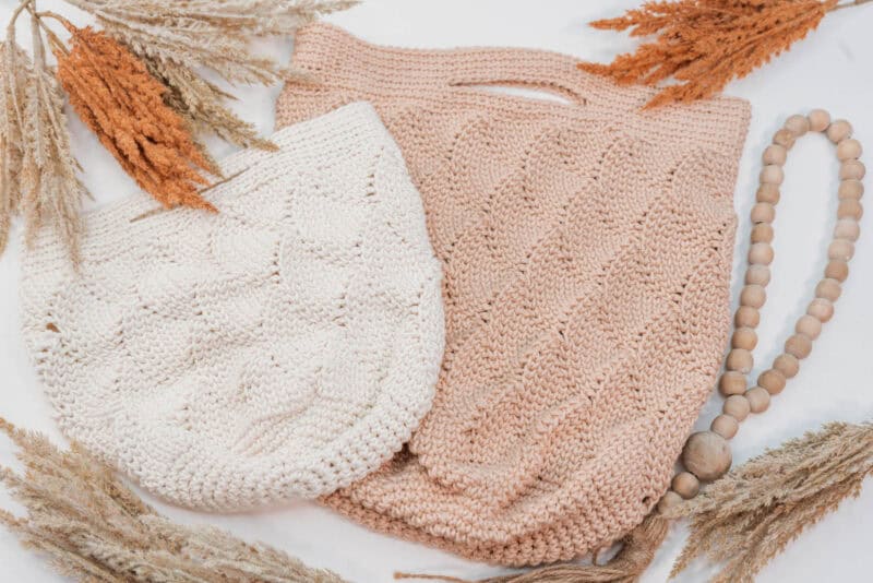 Two knitted bags, one cream and one beige, lie flat on a surface surrounded by dry foliage and a wooden bead garland, reminiscent of a serene journey under the sea.