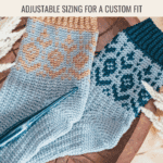 Two pairs of knitted socks with colorwork patterns in blue and tan yarns, displayed on a wooden surface alongside a crochet hook and decorative plants—perfect inspiration for your next Woodland Walker Crochet Socks project.
