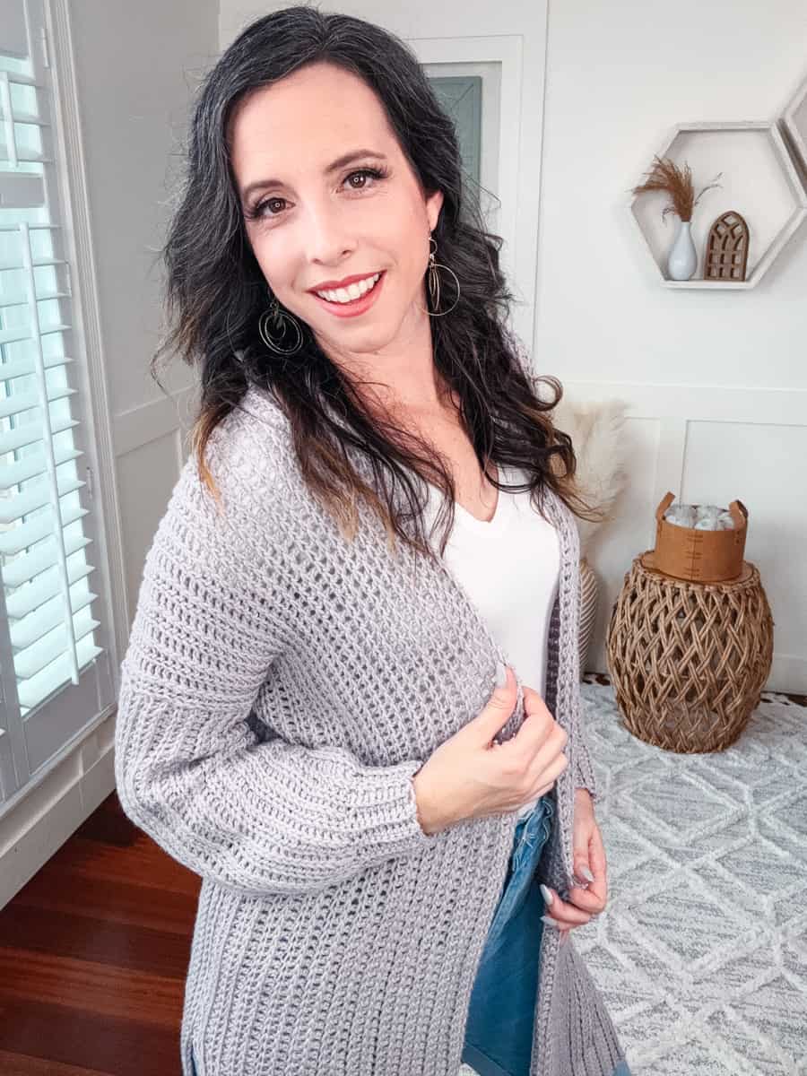A woman with long dark hair wearing a white top, Campside Cardi, and jeans, smiling in a well-lit room with a decorative interior.