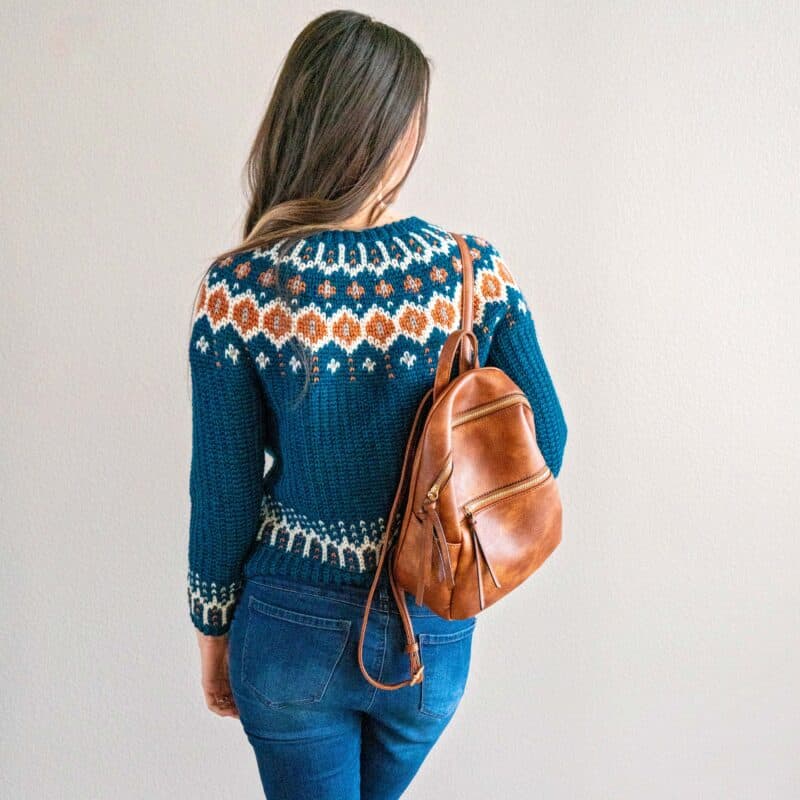 A person with long hair is facing away, wearing a blue and orange Fawning Over You Crochet Sweater, blue jeans, and a brown leather backpack.