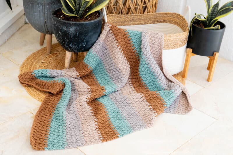 crochet blanket being displayed in a basket surrounded by plants