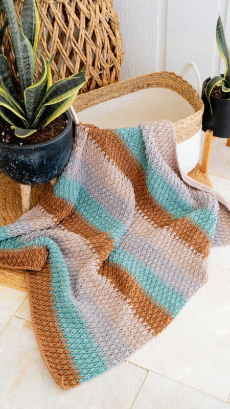 crochet blanket being displayed in a basket surrounded by plants