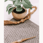 A crocheted square featuring the Alpine Stitch is displayed with a wooden crochet hook, a potted plant, and wooden beads on a neutral surface. Text highlights an Alpine Stitch tutorial and free pattern.