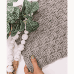 A hand holds a piece of gray crocheted fabric with a basketweave stitch pattern, next to a plant and white beads. Text reads: Basketweave Stitch Tutorial & Free Pattern—plus tips for combining with the Alpine Stitch.