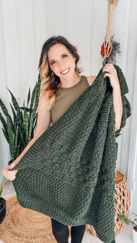 A woman holding a textured green knitted blanket in front of a potted plant.