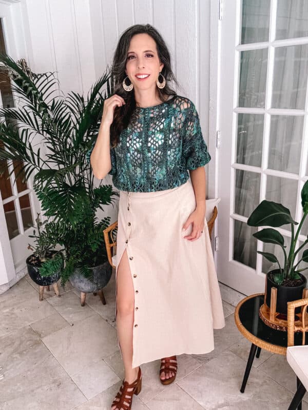 A woman in a teal Key Largo crochet pattern top and beige skirt smiles in a bright room with potted plants.