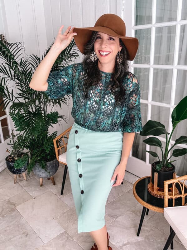 A woman in a Key Largo crochet pattern teal top and mint skirt smiles brightly, tipping her wide-brimmed brown hat, inside a stylish room with green plants.
