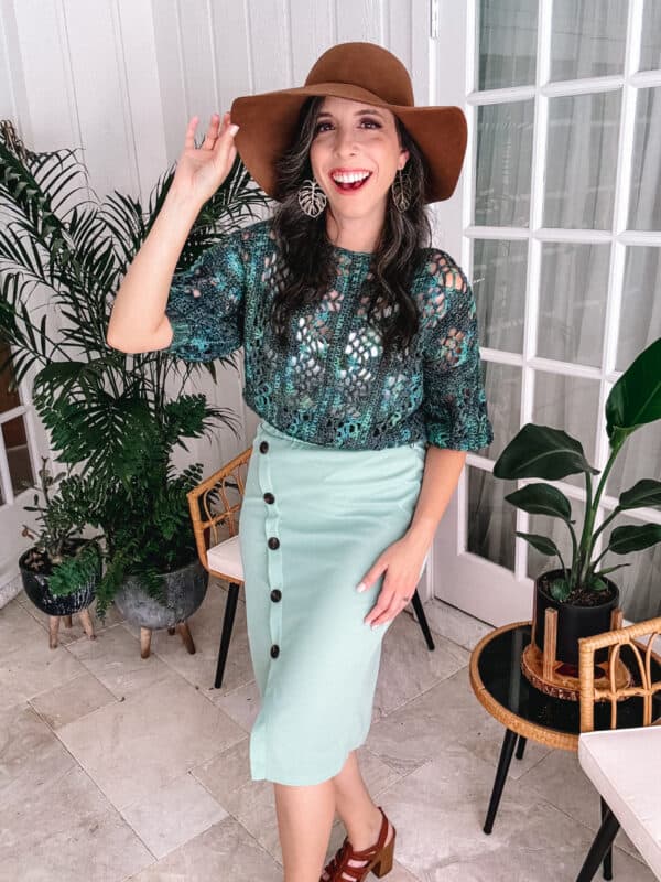 A woman wearing a brown hat, green lace top resembling the Key Largo Top Crochet Pattern, and a mint green skirt with black buttons poses indoors near plants.