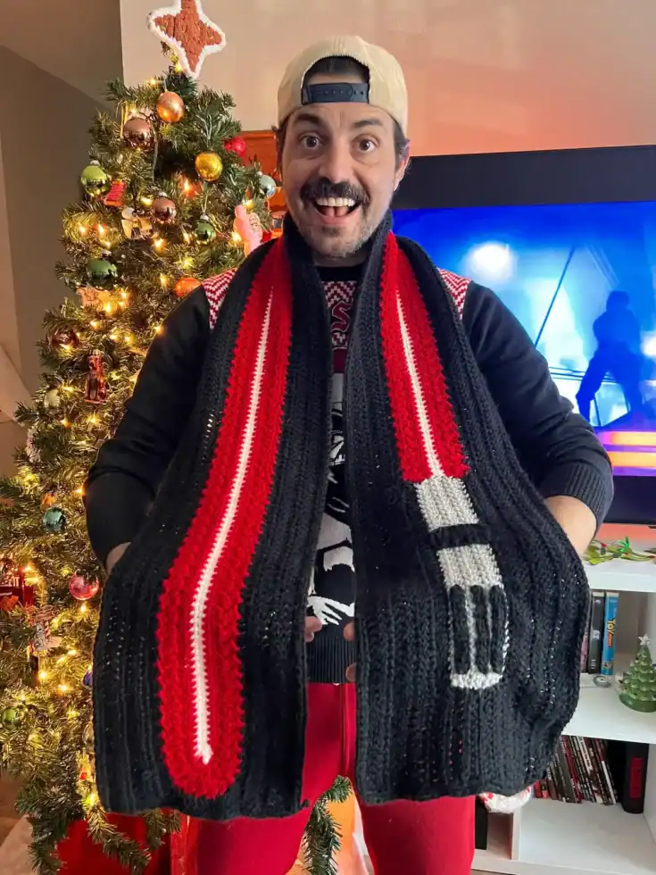 A man wearing a backwards cap and holiday sweater stands in front of a decorated Christmas tree, smiling and holding up a large, red and black Star Wars Knit & Crochet scarf.