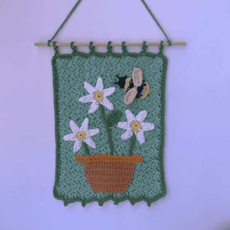 A crochet wall hanging featuring daffodils and a bee.
