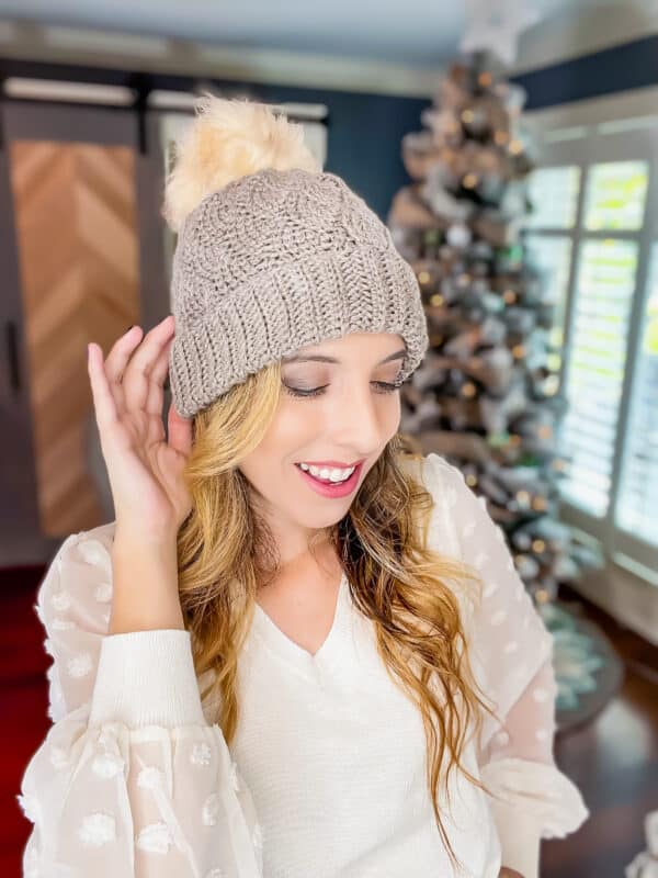 A woman wearing a beanie with a pom pom in front of a christmas tree.