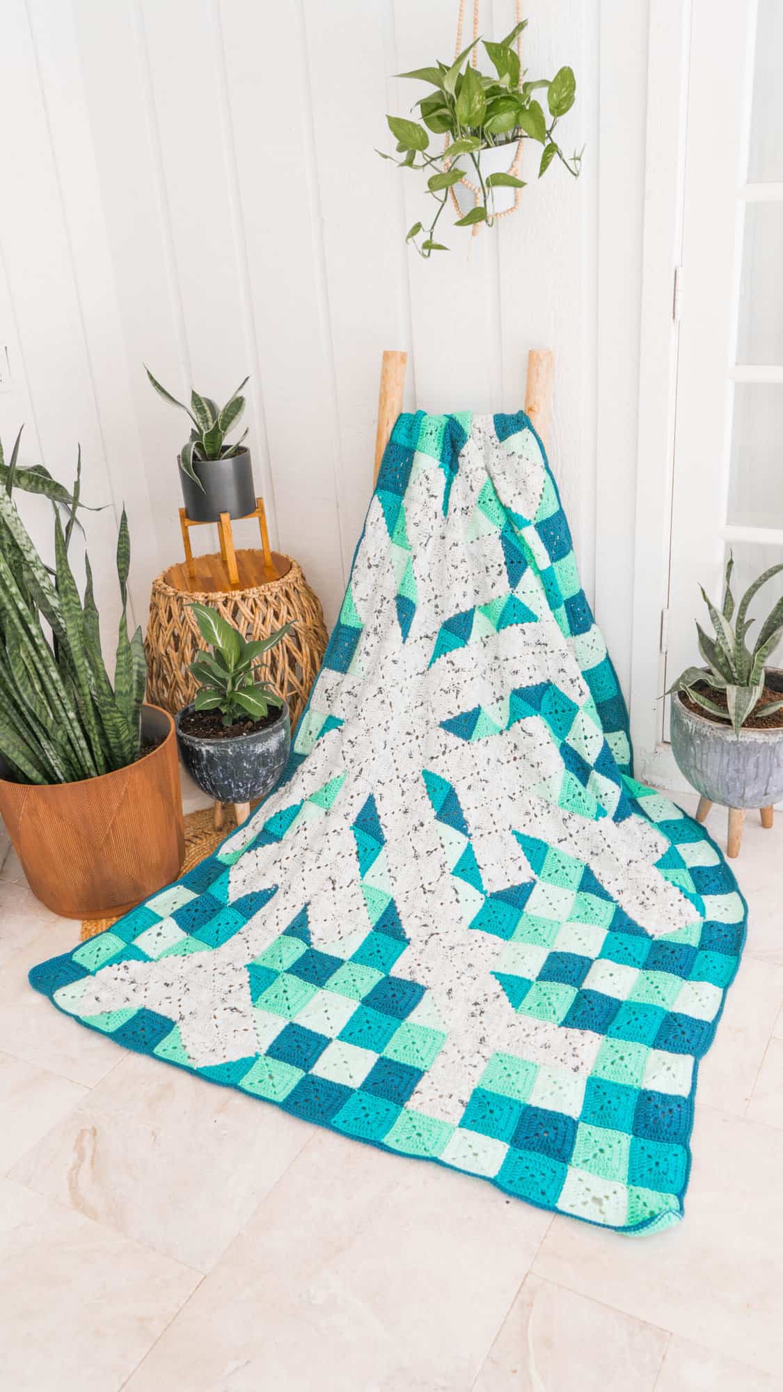 A granny square blanket sitting on a floor next to a potted plant.