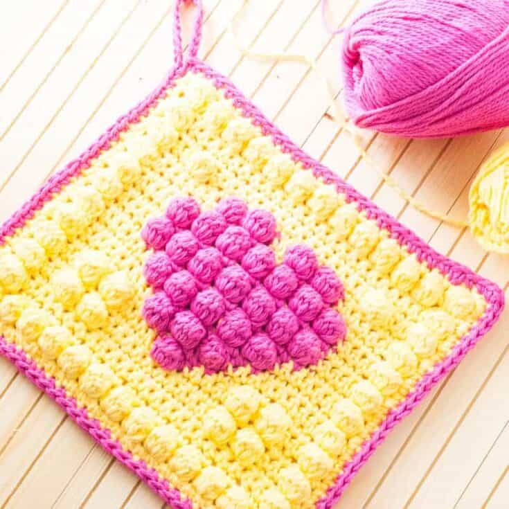 A yellow and pink crocheted heart shaped dishcloth on a wooden table.