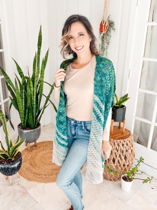 A woman wearing jeans and a crocheted cardigan standing in front of a potted plant, showcasing her elegant On The Bias Crochet Shawl.