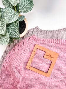 A pink crochet sweater stacked on top of a brown crochet sweater with a gauge tool and green plant.