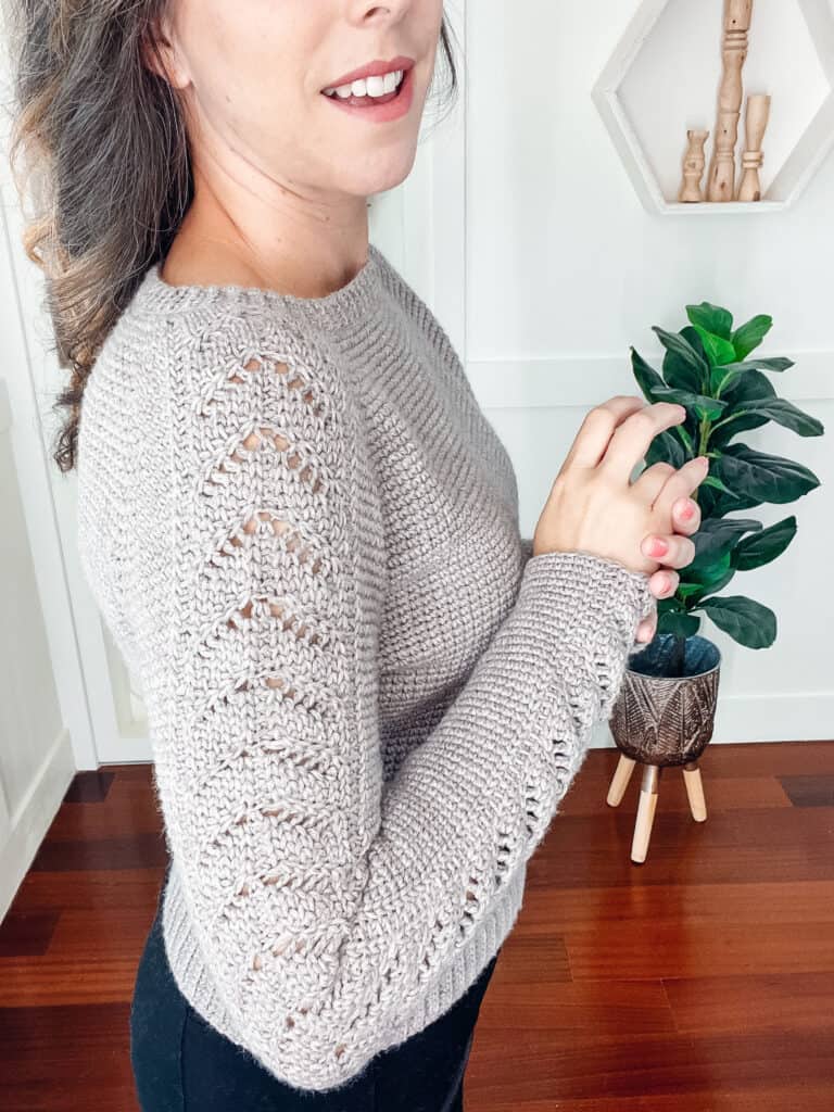 A woman wearing a brownish grey crochet sweater and tan pants standing indoors with a plant and decor behind her