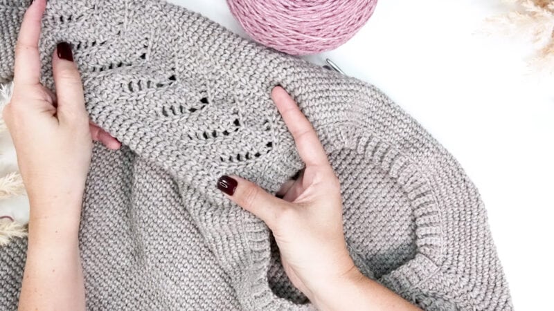 Hands hold a Crochet Lace Sleeve Sweater in beige; its delicate pattern stands out as a ball of pink yarn and a crochet hook rest nearby on the white surface.