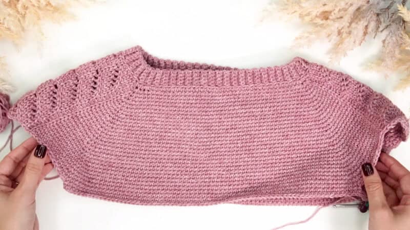 A pair of hands holds a partially finished pink Crochet Lace Sleeve Sweater on a white surface, with decorative dried plants at the corners.
