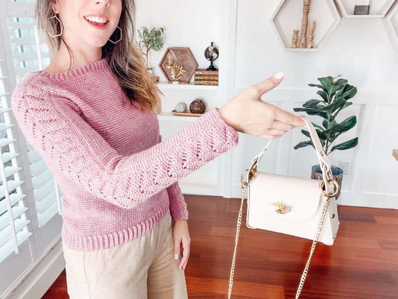 Woman wearing a pink crochet lace sleeve sweater and beige pants holds out a white handbag with gold chain straps in a bright, modern room.