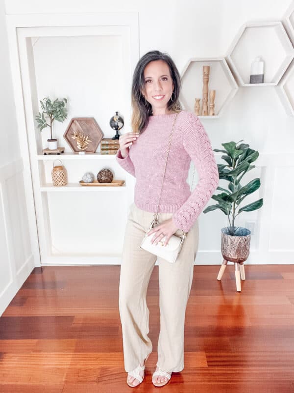 A woman sweating a pink crochet sweater and tan pants standing indoors with a plant and decor behind her