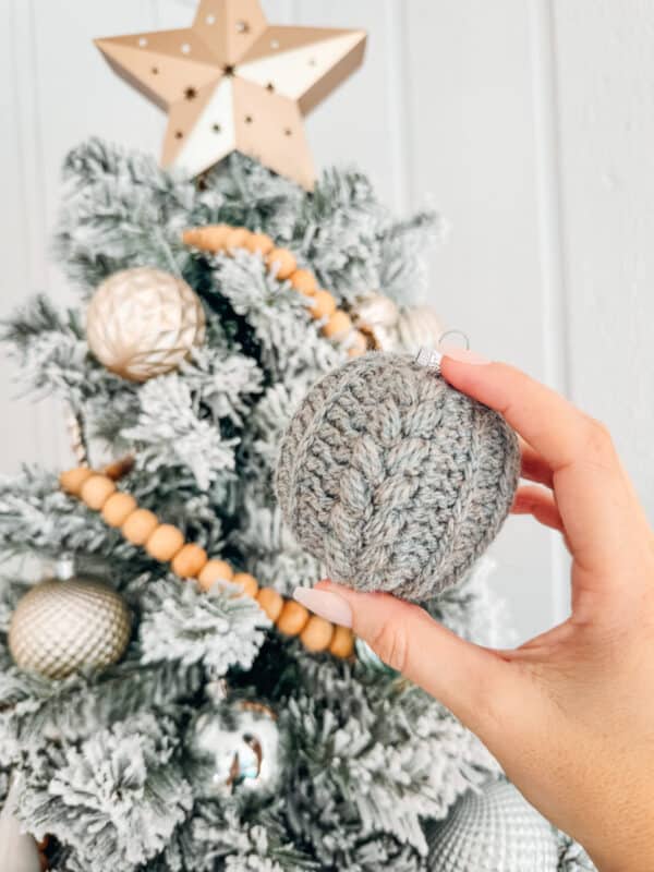 A person holding a crocheted christmas ball in front of a christmas tree, showcasing their crochet skills.