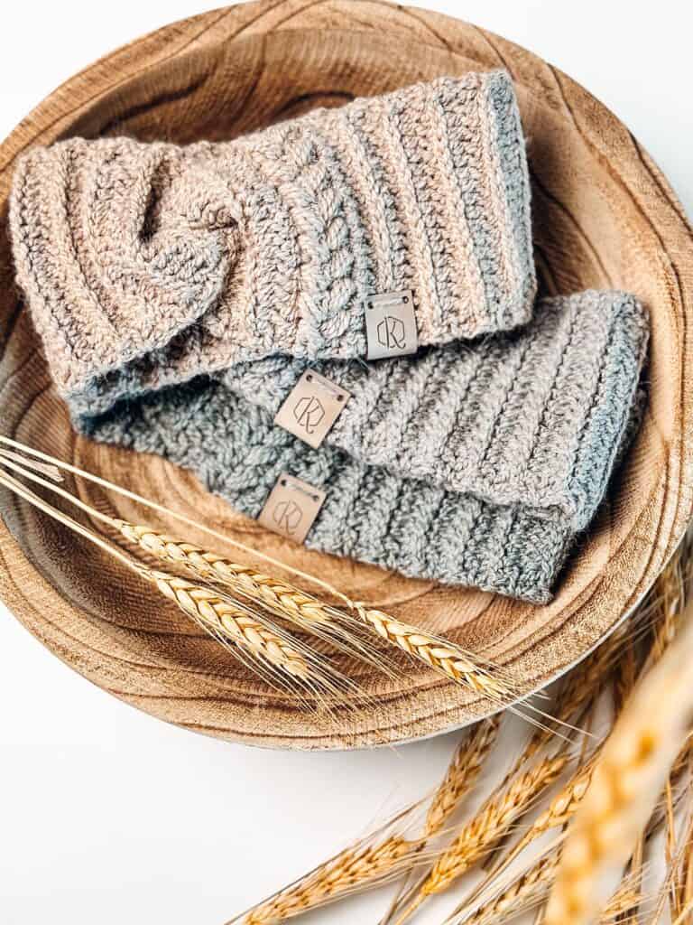 Two crocheted mittens and a crochet ear warmer sitting on a wooden bowl.