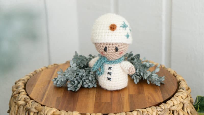 A crocheted snowman wearing a crochet granny square cardigan, sitting on top of a wicker basket.