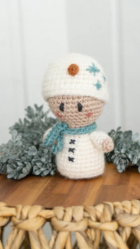 A crocheted snowman wearing a crochet granny square cardigan sitting on a wooden table.