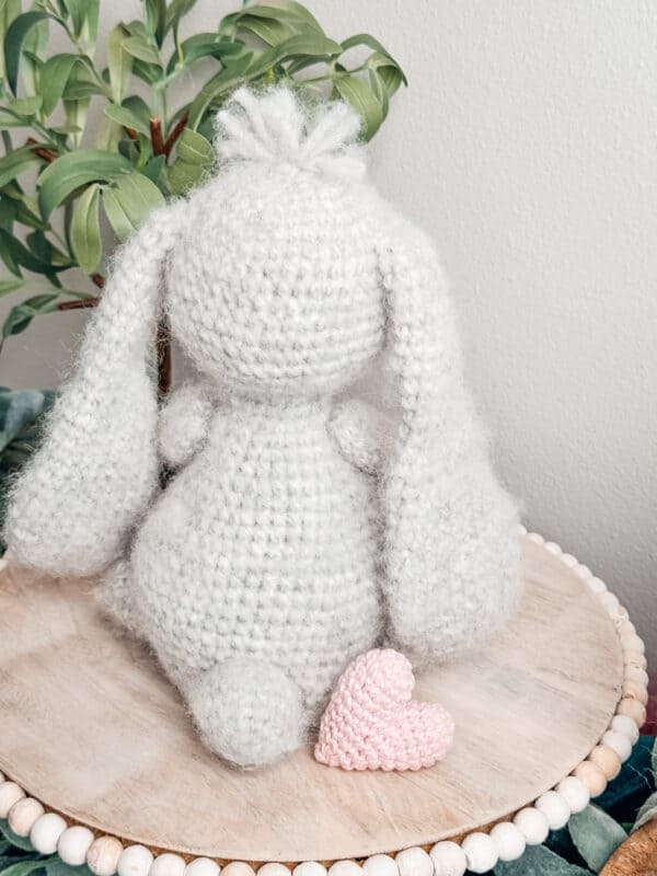 A crocheted bunny sitting on top of a wooden plate, sporting a cute crochet granny square cardigan.